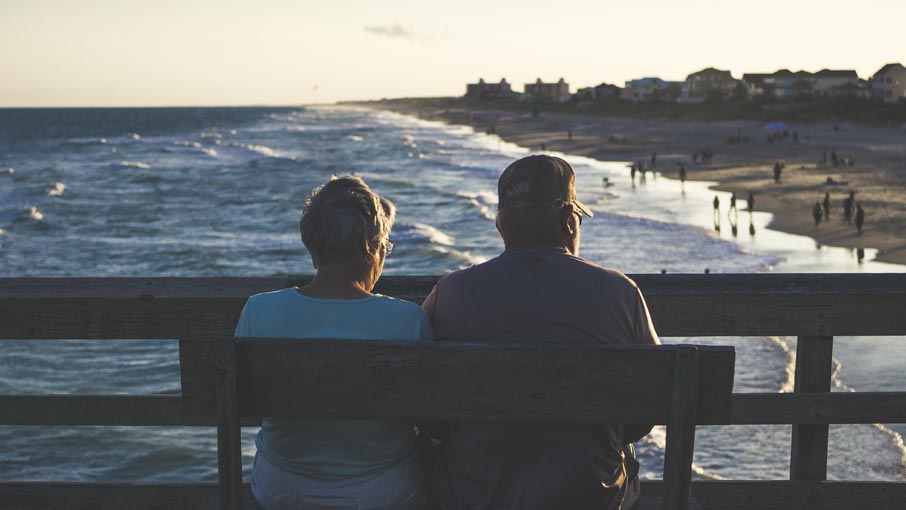 A couple on a pier, sitting on a bench, overlooking the beach.